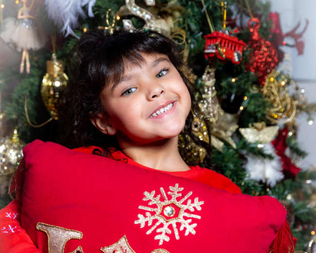 A girl smiles while holding a red decorative pillow in front of a Christmas tree adorned with ornaments and lights.
