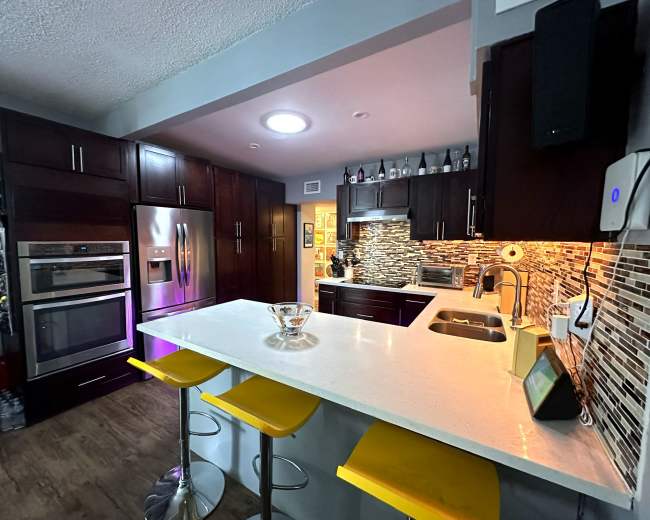 The kitchen features dark wood cabinets, stainless steel appliances, a white countertop, and yellow bar stools at a breakfast island.