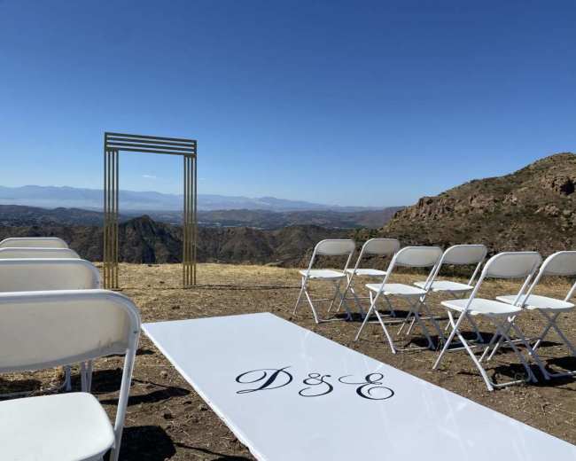 A wedding setup with white chairs and a decorated aisle leading to a gold arch overlooking a mountainous landscape.