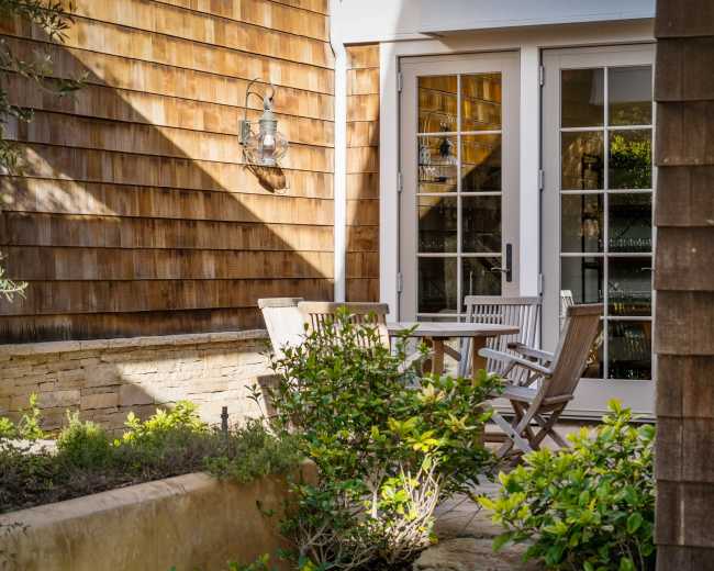 A wooden patio with a small table and chairs is set beside two buildings with textured brown siding.