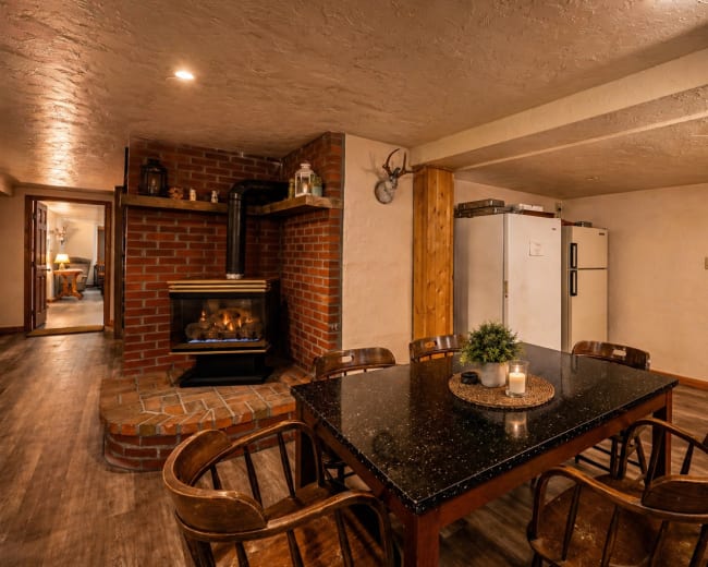 A dining area features a dark granite table surrounded by wooden chairs, with a brick fireplace and a white refrigerator in the background.