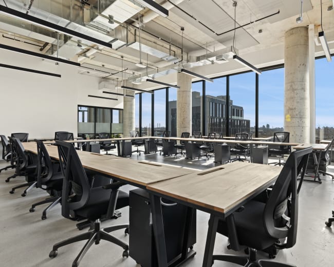 The image shows a modern office space with multiple large desks arranged in a U-shape, surrounded by black ergonomic chairs and large windows allowing plenty of natural light.