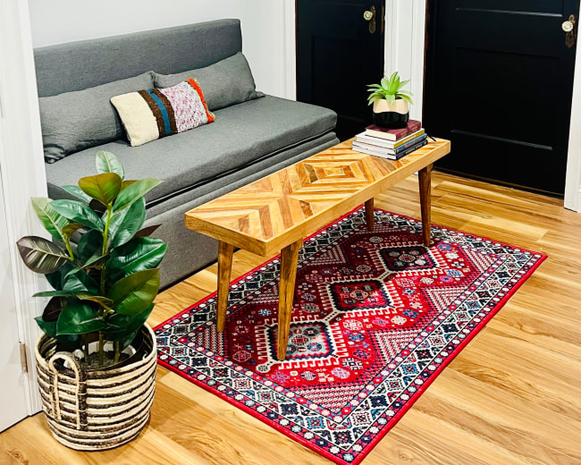 The image shows a cozy living room area featuring a gray sofa, a patterned rug, a wooden coffee table, and a plant beside two closed black doors and a wall art of a cow's face.