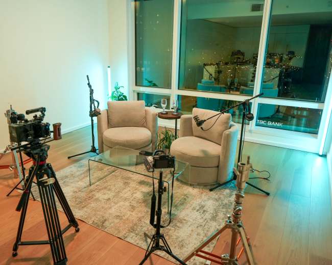 The image shows a modern sitting area with two light gray armchairs, a glass coffee table, and various camera equipment set up around it.
