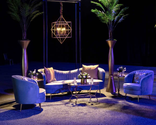 The image shows a stylish seating arrangement with a curved sofa, two armchairs, and decorative tables, set beneath a geometric light fixture and flanked by tall potted plants.