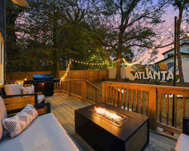 The image shows a wooden deck with modern seating and a fire pit, surrounded by trees and string lights, with a mural that reads "ATLANTA" on a nearby wall.