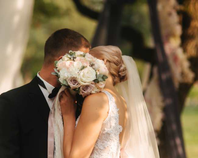 A bride and groom share a kiss while the bride holds a bouquet of roses, standing in front of a decorated outdoor ceremony backdrop.