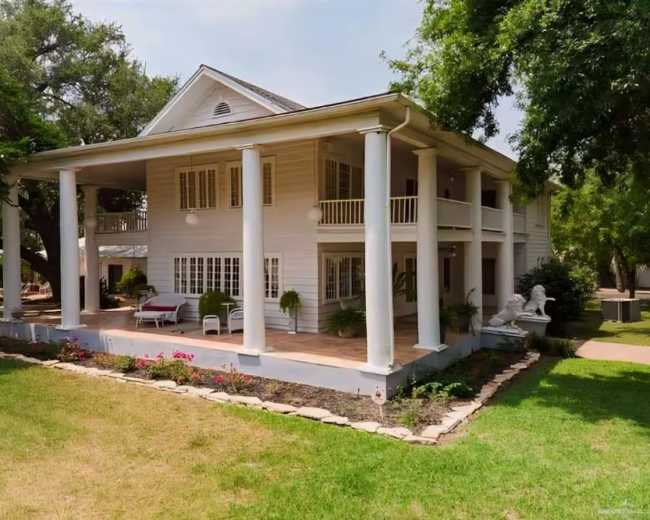 The image shows a two-story, white wooden house with large columns, surrounded by a green lawn and landscaped flowerbeds.