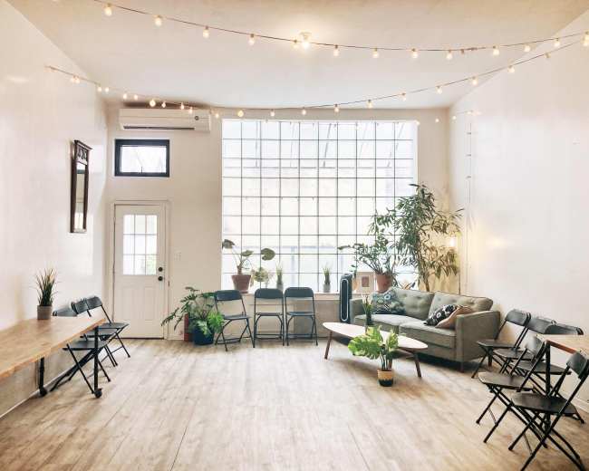 The room features large windows, a cozy sofa, several plants, and multiple folding chairs arranged around two tables.