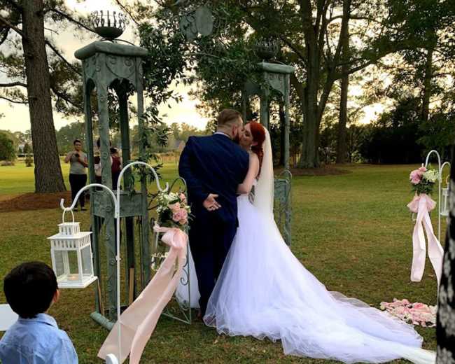 A couple shares a kiss under an ornate archway adorned with flowers during their outdoor wedding ceremony, while a child watches nearby.