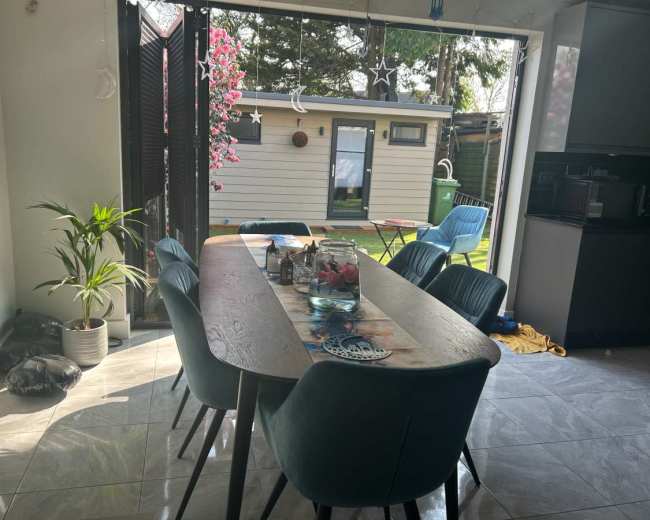 A dining table with blue chairs is set in a brightly lit kitchen, facing an outdoor area featuring a small building and greenery.