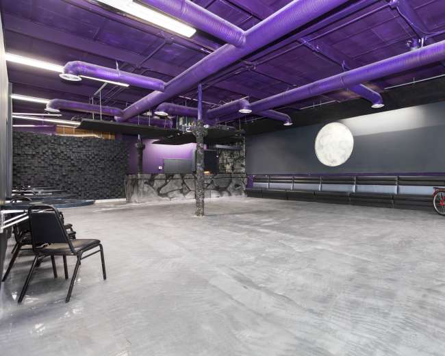 The image shows a spacious, purple-themed room with concrete flooring, featuring black chairs, a decorative bike, and a large wall with a moon mural.