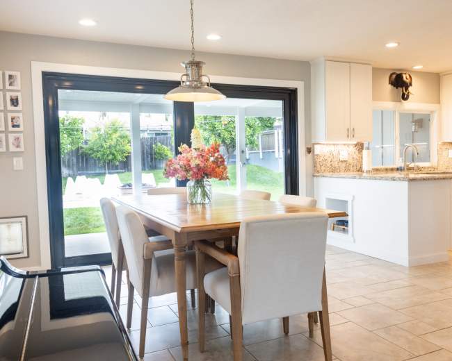 A modern dining area features a wooden table with floral centerpieces, surrounded by light-colored chairs, adjacent to a kitchen with white cabinets and granite countertops.