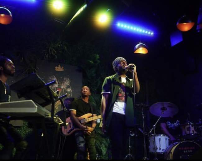 A band performs on stage at Rich & Pour, featuring a vocalist, keyboardist, guitarist, and drummer under colorful lights.