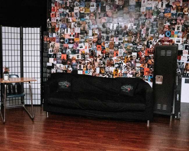 The image shows a room with a black couch against a wall covered in colorful posters, a wooden table with a chair nearby, and a metal locker.
