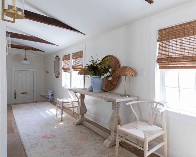 The image features a well-lit hallway with white walls, a wooden table, two chairs, and decorative elements including a vase and a rug.