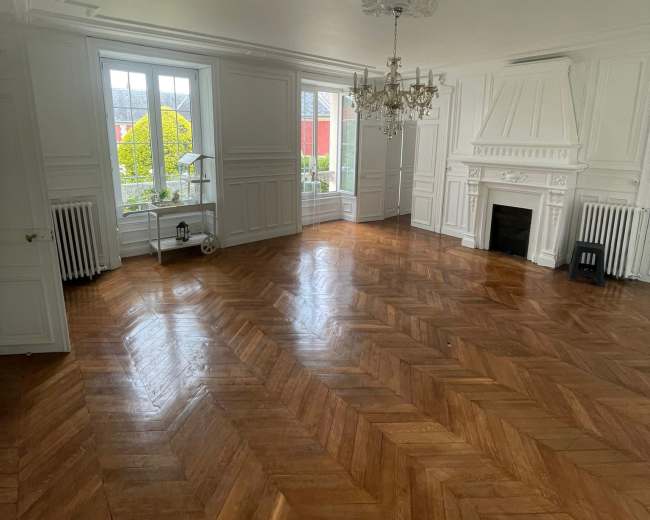 A well-lit, empty room with herringbone wooden flooring, white paneled walls, and a chandelier hanging from the ceiling.