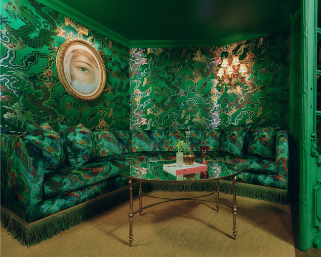 The image shows a vibrant green lounge area with an ornate round couch, a glass table, and a painting on the wall.