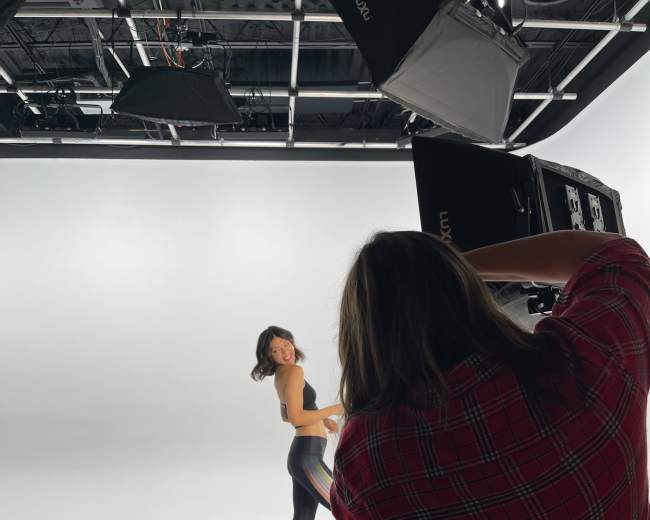 A woman in athletic wear poses while another person adjusts a lighting setup in a photography studio.