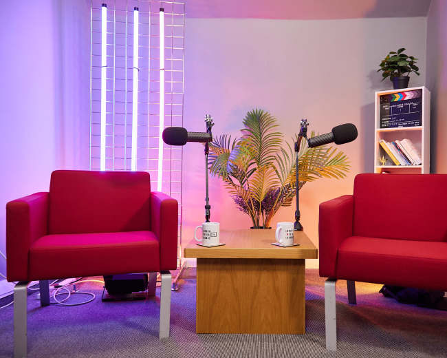A small interview setup featuring two red chairs facing a wooden table, with microphones mounted on stands and a backdrop of decorative plants and lighting.