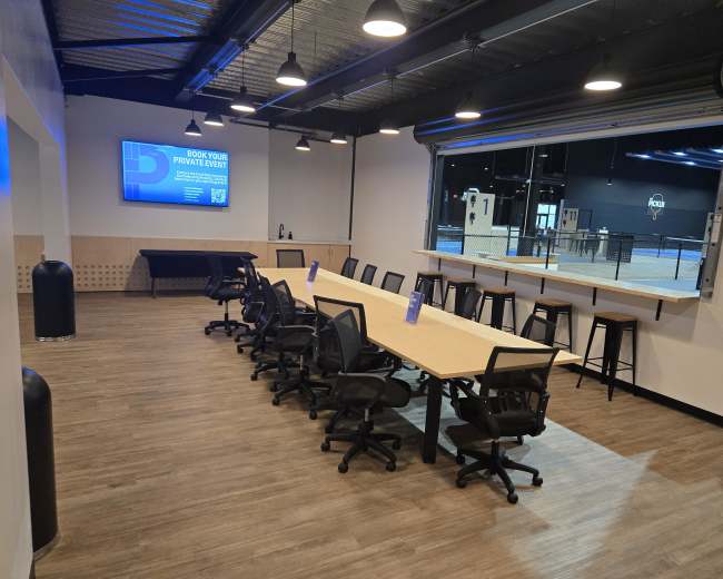 The image shows a modern conference room featuring a long table with multiple ergonomic chairs, a large screen on the wall, and a window overlooking an outdoor area.
