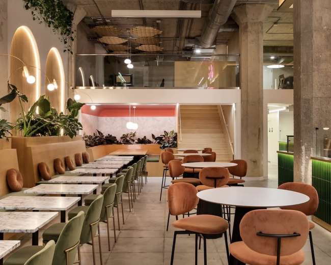 The image shows a modern café interior with tables, colorful seating, and greenery, featuring a stylish staircase and decorative lighting.