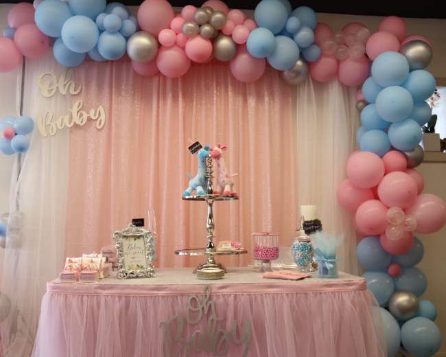 A decorated table with a pink tablecloth features a tiered stand of desserts, surrounded by a balloon arch in pastel colors and a sign reading "Oh Baby."
