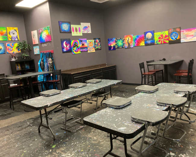 The image shows an art studio with several tables and chairs, surrounded by colorful artwork displayed on the walls.