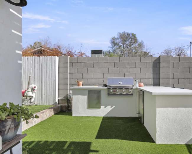The image shows an outdoor kitchen area featuring a built-in grill, a countertop, and artificial grass in a fenced yard.