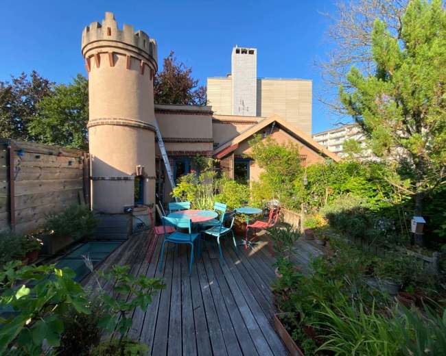 The image shows a rooftop garden featuring a turreted structure, surrounded by lush greenery and colorful outdoor furniture.