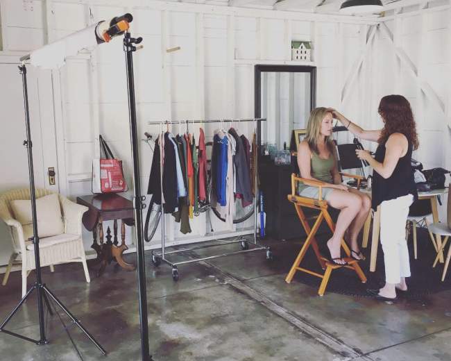A woman sits in a makeup chair while another woman applies makeup in a well-lit studio with a clothing rack and various items in the background.