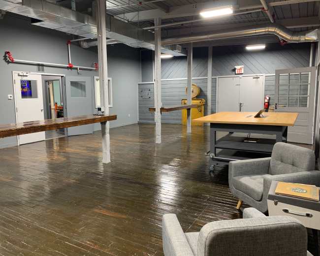The image shows an empty office space with wooden floors, several tables against the walls, and a seating area with a chair and side table.