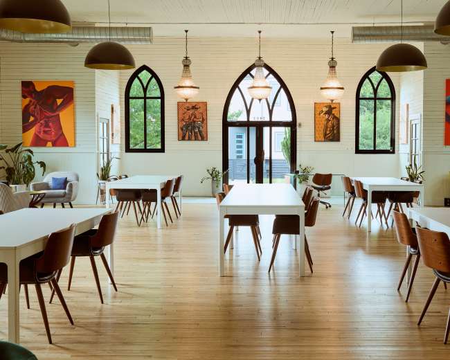 The image shows a spacious, well-lit dining area with white tables and wooden chairs, featuring large windows and colorful artwork on the walls.
