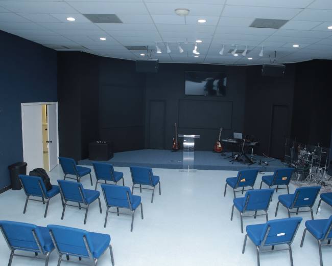 A sparsely furnished performance space with blue chairs arranged in rows facing a stage area that includes musical instruments and a podium.