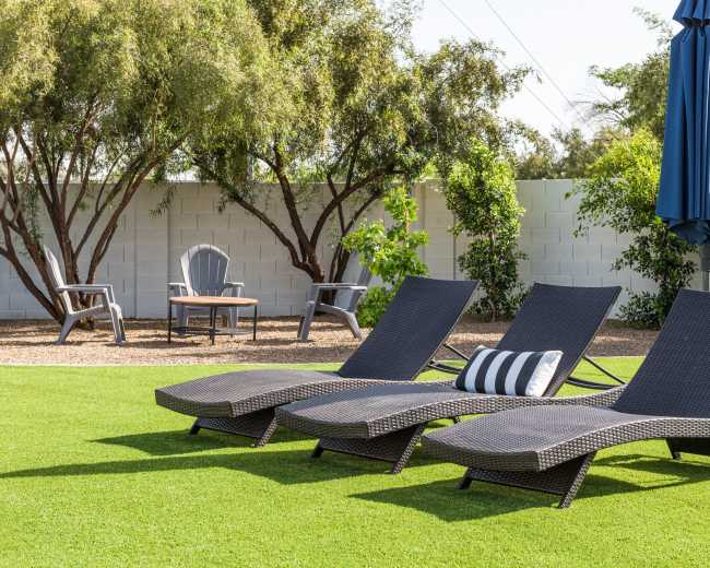 Three lounge chairs are positioned on a green lawn with a blue umbrella and seating area in the background.
