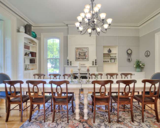 A long, white dining table surrounded by wooden chairs is set in a well-lit room with large windows and decorative shelving.
