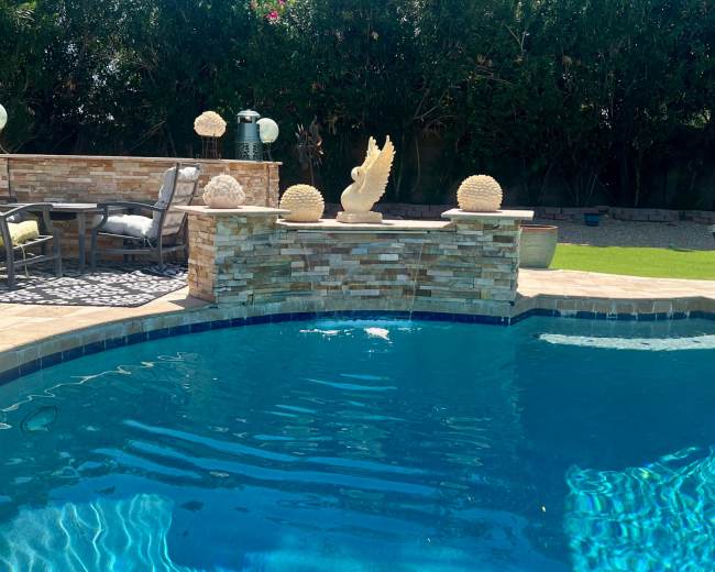 A pool surrounded by stone features and outdoor furniture, with decorative objects and lush green foliage in the background.