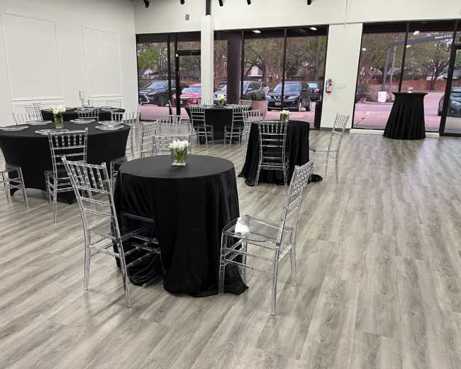 The image shows an event space with several black-covered round tables, each surrounded by clear chiavari chairs, arranged in a well-lit area with large windows.