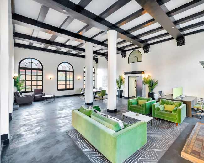 The image shows a spacious interior with high ceilings, featuring green sofas, large windows, and an ornate fireplace.
