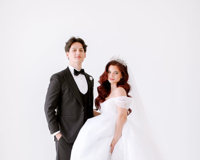 A couple stands together, the woman wearing a white wedding gown and tiara, while the man is dressed in a black tuxedo.