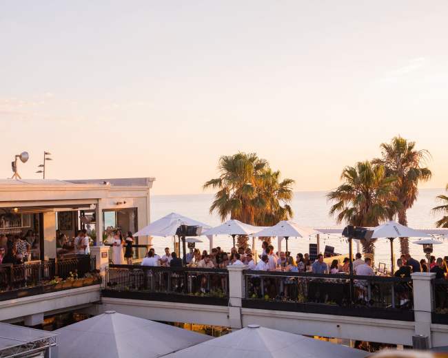 A lively outdoor restaurant overlooking the ocean features patrons enjoying drinks under umbrellas as the sun sets.