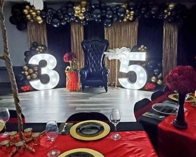 The scene shows a decorated event space featuring a black throne, gold and black balloons, large illuminated numbers "3" and "5," and elegantly set tables with red tablecloths and gold accents.