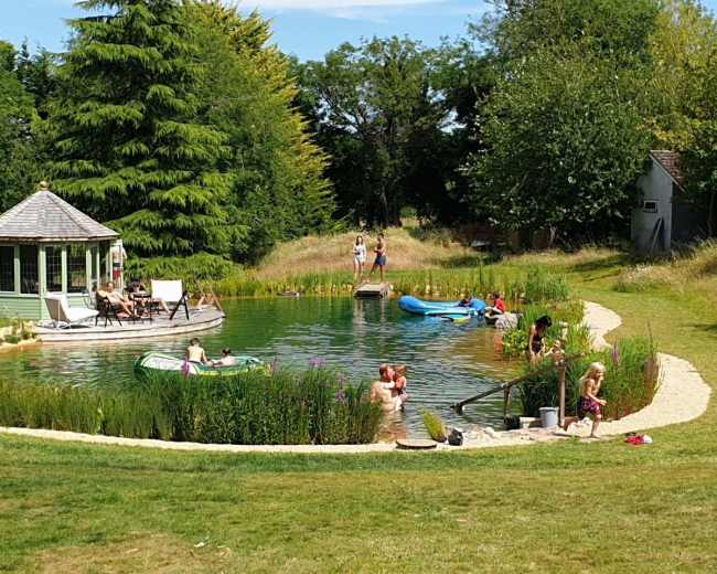 People enjoy a sunny day by a natural swimming pond surrounded by greenery and a gazebo.