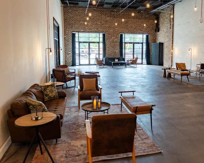 The image shows a spacious, well-lit room with modern seating arrangements, large windows, and an industrial brick wall.