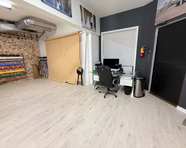 The image shows an office space with a desk and computer, a large window, and a backdrop setup against a wall, along with various photography equipment stored nearby.