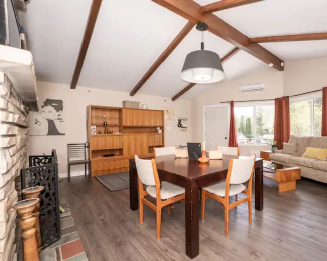 The image shows a spacious living and dining area with a wooden dining table, a sofa, and large windows allowing natural light to enter.