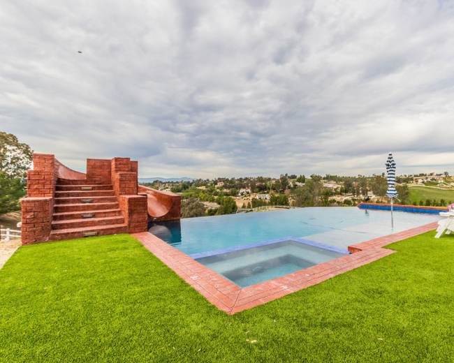 The image shows a large swimming pool with steps leading down to it, surrounded by green grass and overlooking a landscape of houses and trees under a cloudy sky.
