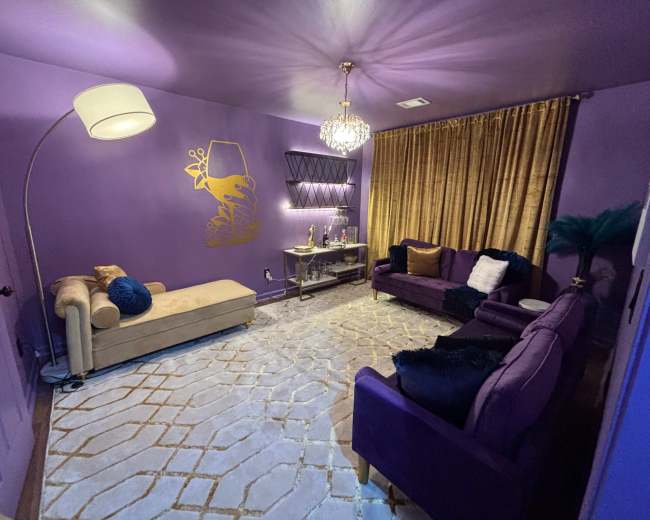 The image shows a purple-themed living room with a chaise lounge, a coffee table, two sofas, a large rug, and gold curtains.