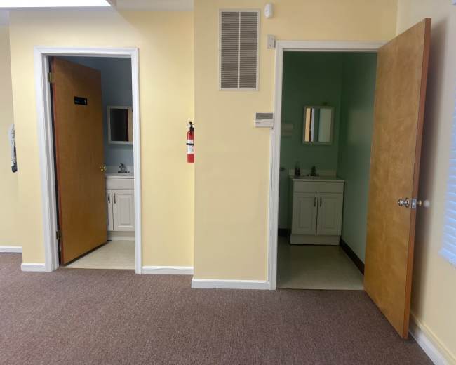 The image shows a hallway with two wooden doors, each leading to small bathroom areas, with one door slightly ajar and the walls painted a light yellow.