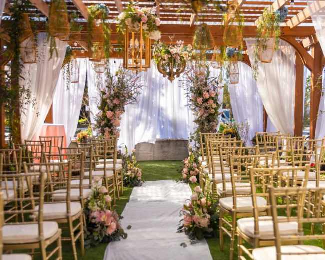 The image shows a wedding ceremony setup with rows of gold chairs facing a floral arch and decorative elements under a wooden pergola.